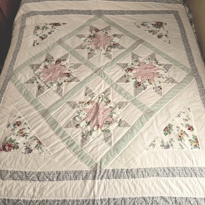 May include: A white quilt with a floral pattern and star-shaped designs. The quilt features pink and gray stars, green striped accents, and floral borders. The overall design is a mix of floral and geometric patterns.