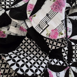 May include: Close-up of a quilt with a patchwork design. The quilt features black and white geometric patterns, along with sections of white fabric printed with musical notes and pink roses. The quilt has a textured appearance.