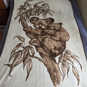 May include: A beige blanket featuring a brown illustration of two koalas in a tree. The larger koala is embracing a smaller one. The tree and leaves are also brown, set against a neutral background.