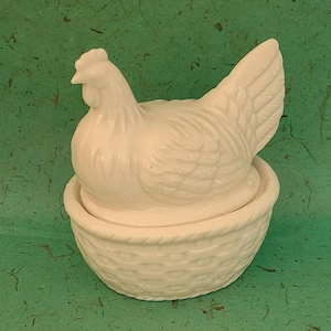 May include: White ceramic chicken figurine sitting in a white ceramic basket. The chicken has a detailed design and is sitting on a nest.