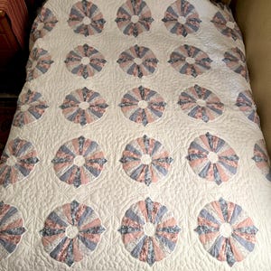 May include: A bed quilt featuring a repeating pattern of circular floral motifs in shades of dusty pink, slate blue, and white. The quilt has a cream-colored background with intricate quilted stitching and a scalloped border edge in a floral print.