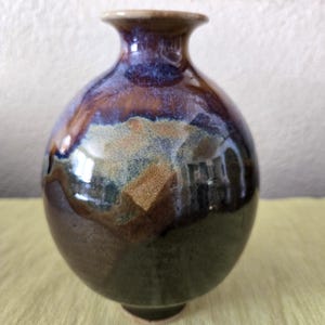 May include: A handcrafted ceramic vase with a rounded body and a narrow neck. The vase features a glossy glaze in shades of brown, blue, and green, creating a unique and artistic appearance. The vase is approximately 6 inches tall.