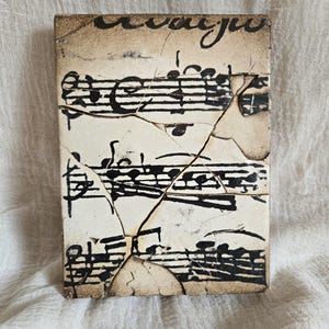 Könnte beinhalten: Rechteckige Keramikfliese mit einem Vintage-Notenmuster. Die Fliese zeigt schwarze Noten und Notenlinien auf cremefarbenem Hintergrund. Die Fliese hat ein rissiges, gealtertes Aussehen mit sichtbaren Rissen und einer verwitterten Ästhetik.