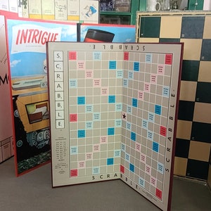 May include: A collection of vintage board games, including Scrabble, Intrigue, and Monopoly. The Scrabble board is open, displaying the game grid with letter and score spaces. Other game boxes are visible in the background, suggesting a game night theme.