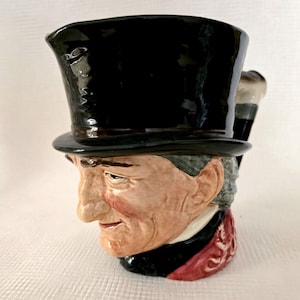 Peut inclure: Une cruche en céramique en forme de personnage portant un haut-de-forme noir. La figurine a un teint clair, des cheveux gris et un col à motifs rouges. L'anse de la cruche est en forme de canne avec un pommeau de couleur argentée.