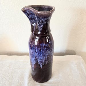 May include: A purple ceramic pitcher with a white glaze. The pitcher has a unique, slightly uneven shape and a wide mouth.
