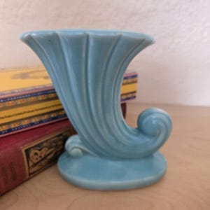 May include: A light blue ceramic vase with a fluted design and a curled handle. The vase is sitting on a wooden surface.