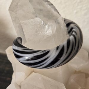 May include: A black and white swirl glass earring. The earring is a curved, circular shape with a glossy finish. The design features alternating black and white stripes. The earring is displayed on a clear, crystalline surface.