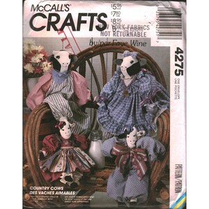Op de afbeelding: McCall's Crafts patroon 4275 door Faye Wine met vier gevulde koe-poppen in diverse outfits. De poppen dragen jurken en shirts van blauwe, roze en bedrukte stoffen. De patroon-envelop bevat tekst en een streepjescode.