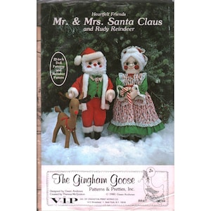 Pode incluir: Um pacote de moldes de costura intitulado Heartfelt Friends Mr. & Mrs. Santa Claus and Rudy Reindeer para uma boneca de 56 cm. A capa mostra um Papai Noel, Mamãe Noel e uma rena de tecido na neve contra um fundo de pinheiro.