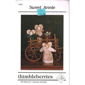 May include: A craft pattern booklet titled Sweet Annie by thimbleberries featuring four fabric dolls with blonde hair. Three dolls are inside a wooden wagon with spoked wheels, and one doll stands in front. The text includes JL-8021 and 205 Jefferson St.