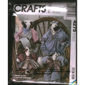 Op de afbeelding: Een McCall's Crafts naaipatroon envelop 4275 door Faye Wine met illustraties van vier gevulde koeienpoppen. De poppen dragen diverse jurken en outfits van stof met patronen. De tekst bevat 'CRAFTS', '4275' en veiligheidswaarschuwingen.