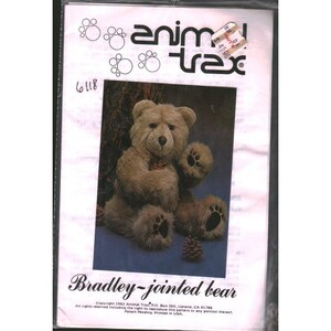 May include: A sewing pattern for a Bradley-jointed bear by Animal Trax. The image shows a plush bear with a light brown fur, sitting on a blue surface. The pattern's packaging includes the text "Animal Trax" and "Bradley-jointed bear".