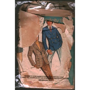 May include: A vintage Simplicity sewing pattern envelope enclosed in a clear plastic bag. The paper cover features illustrations of two outfits: a double-breasted blue blazer with light trousers and a plaid blazer with brown trousers. The paper is torn.