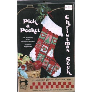 May include: A Christmas stocking kit with the text "Pick a Pocket". The 24-inch stocking is red with white trim and appliqued squares featuring hearts, gingerbread men, and trees. The kit includes 8 surprise pockets and is machine appliqued.