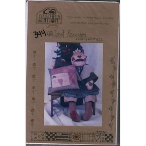 Op de afbeelding: Een bruine papieren verpakking met een country poppenpatroon. Het patroon bevat een pop met een hartvormig gezicht, zittend op een kleine houten bank, met een kussen met een hartontwerp. De tekst "Janet Rosanna" is zichtbaar.