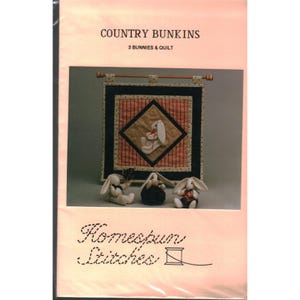 以下が含まれることがあります： COUNTRY BUNKINS 3 BUNNIES & QUILTおよびHomespun Stitchesと題されたクラフトパターン。画像には、中央のダイヤモンドに白いウサギが描かれ、濃い青と赤のチェック柄の縁取りが施された壁掛けキルトと、3体の小さな白いぬいぐるみのウサギが写っています。