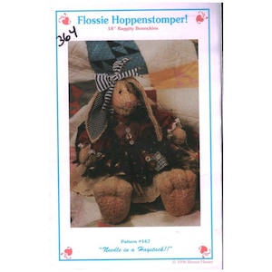 Może przedstawiać: Ręcznie wykonana szmaciana lalka w kształcie królika, o wysokości około 46 cm, z oklapniętymi uszami i paskowaną kokardą. Królik ma na sobie wzorzystą sukienkę z detalami guzików. Obraz zawiera tekst "Flossie Hoppenstomper!" i "Needle in a Haystack!!".
