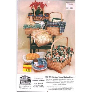 May include: A collection of woven wooden baskets featuring various fabric liners with floral, plaid, and polka dot patterns. Some baskets include ruffles, bows, and lace trim. A rectangular basket holds a loaf of bread, and a small blue basket contains sewing supplies. Text includes RiverTown Warehouse, 6 Brook Road, Hannibal, MO 63401, and R-293 Custom Made Basket Liners.