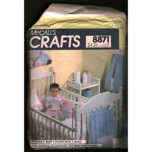 Peut inclure: Une enveloppe de patron McCall's Crafts vintage présentant une chambre de bébé. L'image montre un bébé dans un lit bébé blanc avec une literie rose et un rideau bleu. Le texte de l'enveloppe indique "Décorez la chambre de bébé avec une couette".
