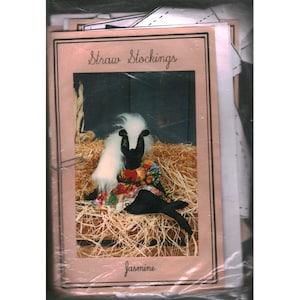 May include: A craft pattern packet titled Straw Stockings featuring a photograph of a black skunk doll with white fur and a floral dress sitting on straw. The packet includes printed instructions and is enclosed in a clear plastic protective sleeve.