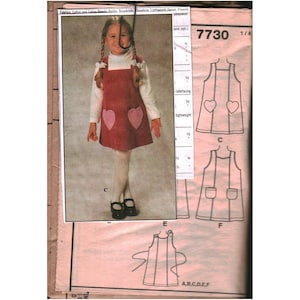 Può includere: Una busta per cartamodello numerata 7730 che mostra la fotografia di un vestito a grembiule con due tasche a forma di cuore indossato sopra una maglia bianca a maniche lunghe. La busta include disegni al tratto di varianti.