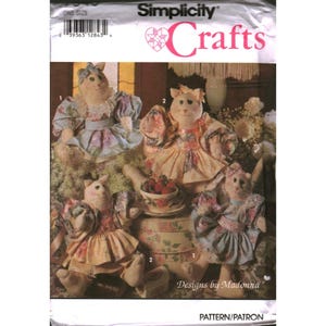 Puede incluir: Un sobre de patrón de costura Simplicity Crafts que muestra cuatro muñecos de gato de tela con vestidos florales, mangas abullonadas y lazos. El paquete incluye el texto Simplicity Crafts y Designs by Madonna, con un código de barras.