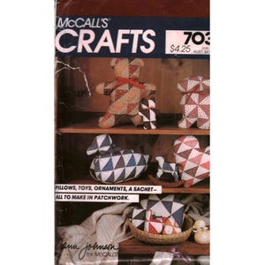 Peut inclure: Couverture du magazine McCall's Crafts avec des créations en patchwork. L'image présente une étagère avec des objets faits à la main, comme un ours en peluche, un canard et un cœur. Le texte indique : "Oreillers, jouets, ornements, un sachet - tous à faire en patchwork."