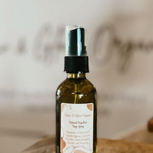 May include: A brown glass spray bottle with a black nozzle and a white label. The label reads "Grow & Glow Organics Natural Repellent Bug Spray" with a list of ingredients. The bottle is on a wooden surface.