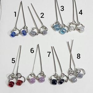 May include: Eight silver hair pins, each with a different colored bead accent. The beads are blue, gray, red, purple, and black. The pins are arranged in a grid pattern.
