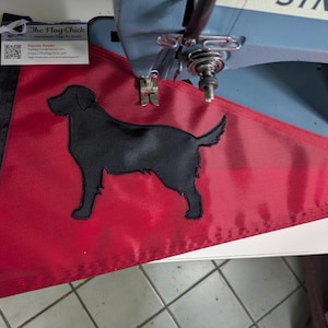 May include: A triangular red flag featuring a black silhouette of a Labrador retriever dog. The flag has a black vertical strip with a brass grommet on the left side. A small white business card with the text The Flag Chick is attached to the top.