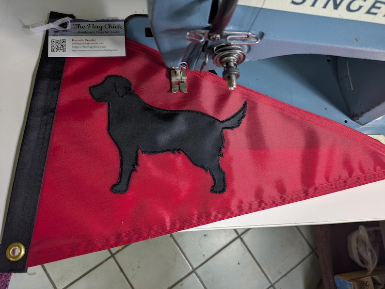 May include: A triangular red flag featuring a black silhouette of a Labrador retriever dog. The flag has a black vertical strip with a brass grommet on the left side. A small white business card with the text The Flag Chick is attached to the top.