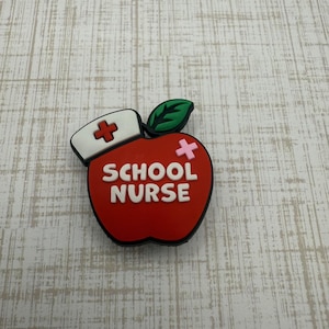May include: A red apple-shaped charm with the words "SCHOOL NURSE" in white. The charm has a green leaf, a white nurse's cap with a red cross, and a pink cross on the apple. The charm is designed for a school nurse.