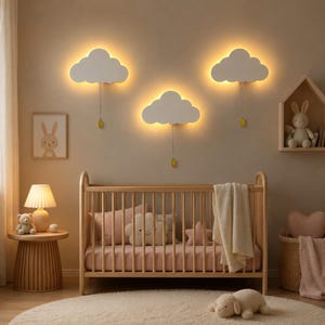 May include: Three white cloud-shaped wall lamps with warm backlighting and hanging pull cords with yellow teardrop-shaped toggles. The lamps are mounted on a beige wall above a wooden crib containing a cream blanket and plush pillows.
