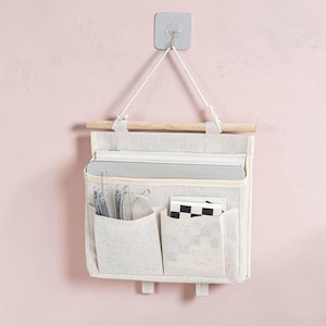 May include: A beige fabric wall hanging organizer with a wooden dowel and white cord. It features one large main compartment and two smaller front pockets containing pens, a clear ruler, glasses, and a small notebook with a black and white grid pattern.