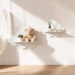 May include: Two white wall-mounted shelves shaped like clouds. The left shelf holds a light brown plush teddy bear and a small green succulent in a white pot. The right shelf holds a glass reed diffuser bottle with black reeds and two small figurines.