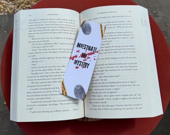 Op de afbeelding: Een bladwijzer met de tekst "INVESTIGATE THE MYSTERY" in een vetgedrukte letter, met een bloedspatterontwerp. De bladwijzer ligt op de pagina's van een boek, met een rode tafel op de achtergrond.