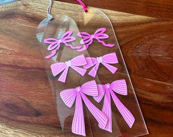 Può includere: Due segnalibri in acrilico trasparente con disegni di nastri rosa. Ogni segnalibro ha un fiocco.