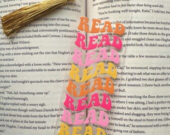 Peut inclure: Un marque-page en acrylique coloré avec le mot "READ" répété cinq fois dans différentes couleurs et styles. Le marque-page est superposé avec chaque mot "READ" se chevauchant légèrement le suivant. Les couleurs sont orange, rose, jaune et rouge.