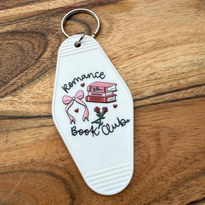 May include: White motel-style keychain with the words "Romance Book Club" and a graphic of books, hearts, a bow, and roses. The keychain has a silver key ring and is on a wooden surface.