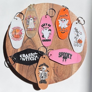 May include: A collection of Halloween-themed motel keychains in various colors, including white, pink, orange, black, and tan. Each keychain features unique graphics and text, such as ghosts, pumpkins, and skeletons. The keychains are arranged on a wooden surface.