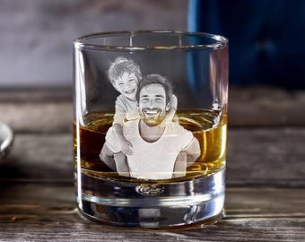 May include: A clear, round whiskey glass filled with amber liquid. The glass features an etched image of a man carrying a child on his back. The image is in black and white, and both figures are smiling. The glass sits on a wooden surface.