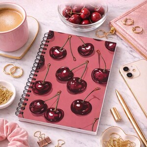 May include: A spiral-bound notebook with a cherry pattern on a pink background. The text "Juicy Cherry Notebook" is at the top. Other items include a pink cup of coffee, a small bowl of cherries, and gold accessories.