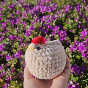 May include: A small, crocheted plush toy shaped like a chicken. The chicken is beige with a red comb and yellow beak. The chicken is sitting in a hand against a background of purple flowers.
