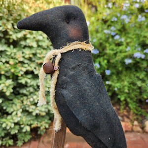 May include: A handcrafted black crow doll with a rustic aesthetic. The crow has a long beak, a jute rope collar, and a small metal bell. The doll is perched on a wooden post against a backdrop of green foliage and blue flowers.