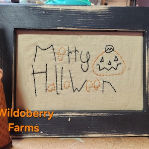 Pode incluir: Uma obra de arte emoldurada com tema de Halloween com as palavras "Merry Halloween" bordadas em fio preto e laranja, juntamente com uma ilustração de abóbora. A moldura preta tem um acabamento envelhecido. O logotipo Wildoberry Farms está na parte inferior.