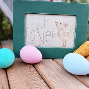 May include: A green wooden frame with the word "Easter" embroidered on a white fabric background. A yellow embroidered chick with a pink bow is in the center of the frame. Three pastel colored eggs and two fabric carrots are on a wooden surface.