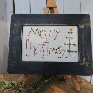 Może przedstawiać: Czarne ramki z haftem na białym materiale z czerwonym haftem, który mówi "Wesołych Świąt" z zielonym i żółtym haftem choinki.