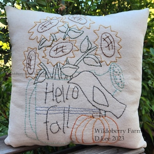 May include: Cream-colored decorative pillow with embroidered fall-themed design. Features sunflowers, pumpkins, and a bird, with the words "Hello fall" stitched in black. The pillow is placed on a wooden surface.