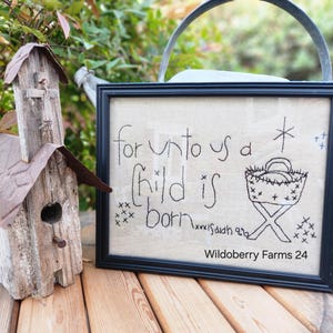 May include: A framed embroidery with the text "for unto us a Child is born" and a star above a manger with a baby inside. The embroidery is signed "Wildoberry Farms 24".